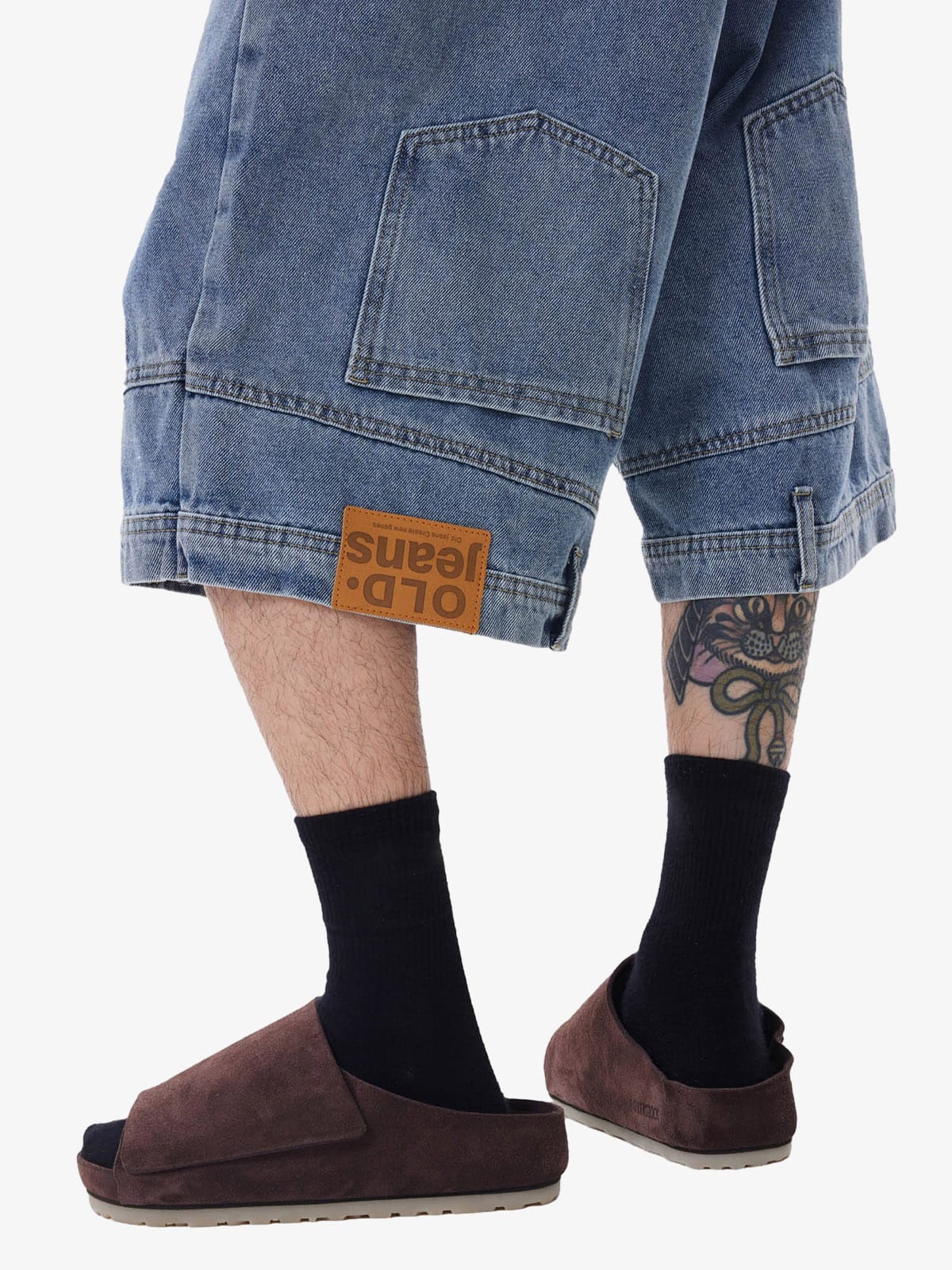 Person wearing blue jeans with a brand label, black socks, and brown shoes on a white background